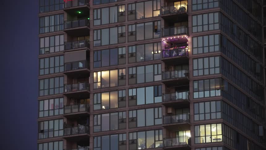 This static midrange shot captures the facade of a modern highrise apartment building in the evening or at nightmany windows and balconies are illuminated from within with one central balcony distinctly decorated with blinking pink or pu