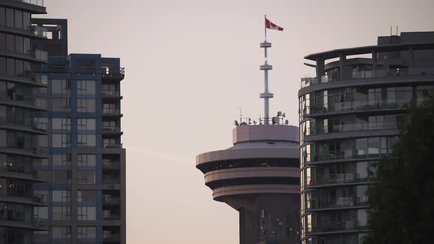 This static midrange shot captures the upper portion of the revolving observation tower of the harbour centre in vancouver flying a canadian flag it is tightly framed between two contrasting modern highrise residential buildings with glass facad
