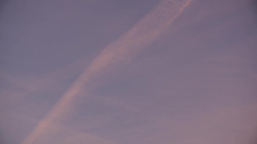 This video shows a wide expanse of sky at sunset or sunrise filled with soft colors of purple pink and pale bluea single long bright contrail vapor trail stretches diagonally across the frame gradually fading as the clip progresses