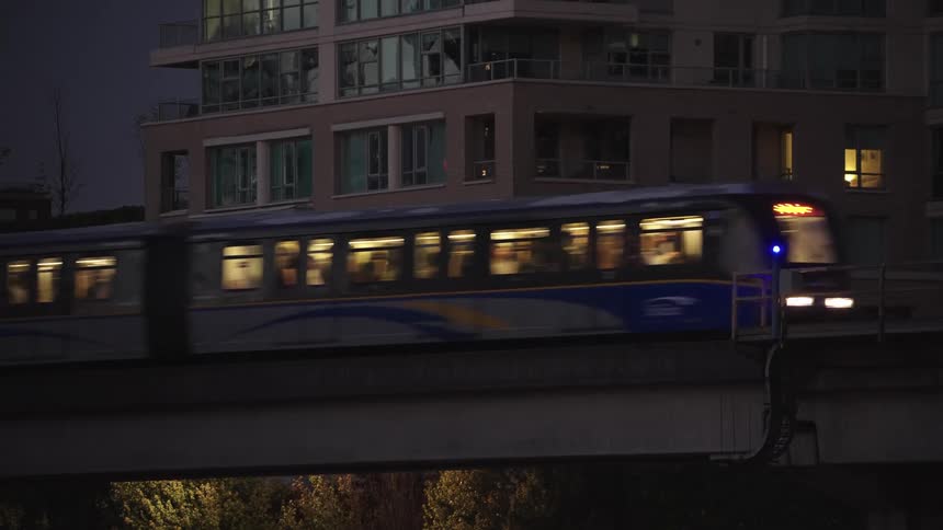 A commuter train speeds across an elevated track in the foreground passing a modern illuminated apartment building at night with a bright blue light signal on the railing