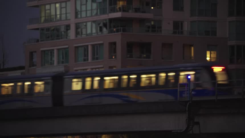 This shot captures the side of a modern apartment building at night with many windows and balconies lit up from withina blue and white commuter train passes quickly from right to left across the lower foreground on an elevated track briefly i