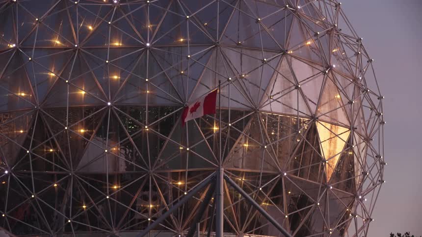 A canadian flag hangs on a pole in front of a large illuminated geodesic dome structure made of steel triangles against a pale evening sky