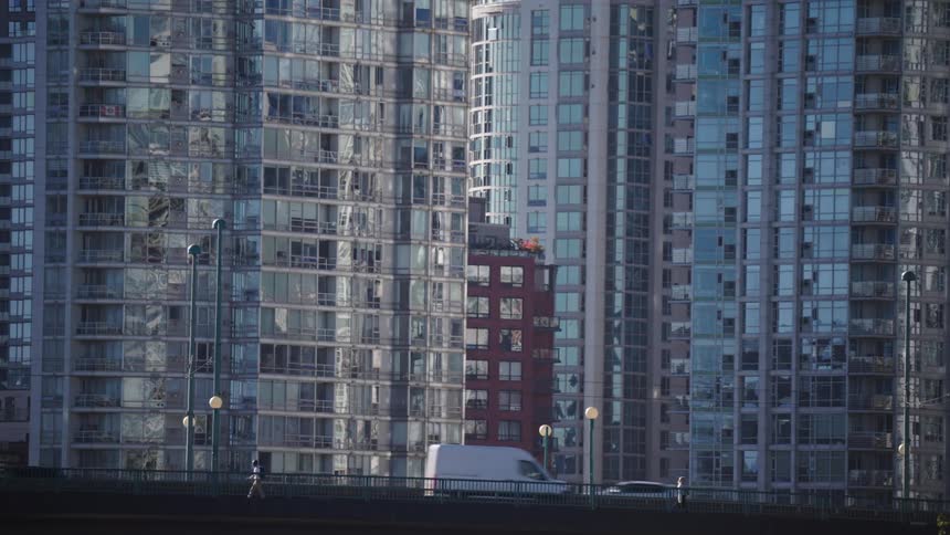 This is a static telephoto shot capturing the vertical facade of two modern highrise apartment towers in bright daylightthe lower foreground features an elevated street or bridge where cars and a small white van pass by creating a strong co