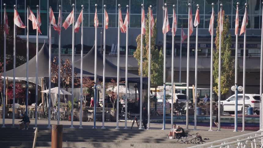 People sit on the steps near a line of canadian flags while others walks and cycle past during the day