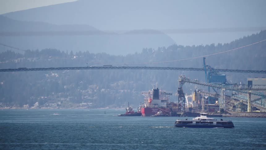 A ferry boat crosses a body of water toward a large industrial port with a red cargo ship docked and a suspension bridge overhead all against a background of hazy forested mountains