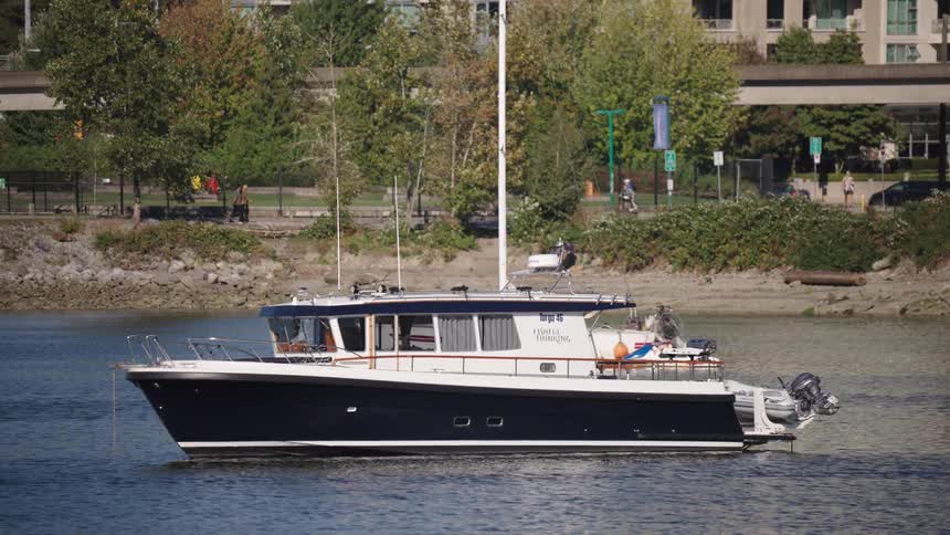 A dark blue and white yacht is anchored in calm water with a lush green tree lined shore in the background