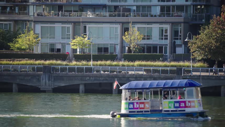 A small blue and white ferry boat with a canadian flag travels lefttoright across a dark river passing a low concrete bridge and modern waterfront apartment buildings on a sunny day