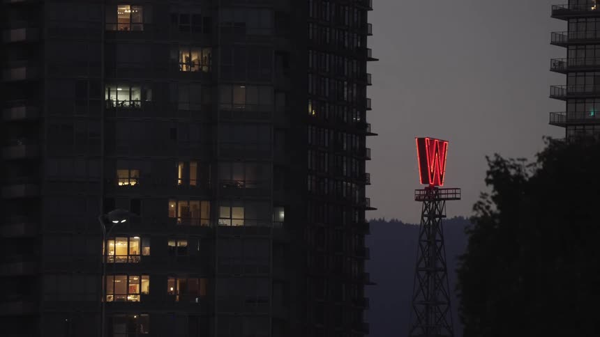 This nighttime or dusk shot captures the facade of a tall dark apartment building with various windows illuminated from withindominating the center of the frame is a large glowing red letter w on a tower which flickers off and then back on