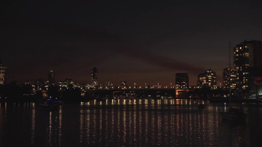 An urban skyline at night with multiple illuminated structures and a central bridge reflected on the dark surface of a body of water in the foreground