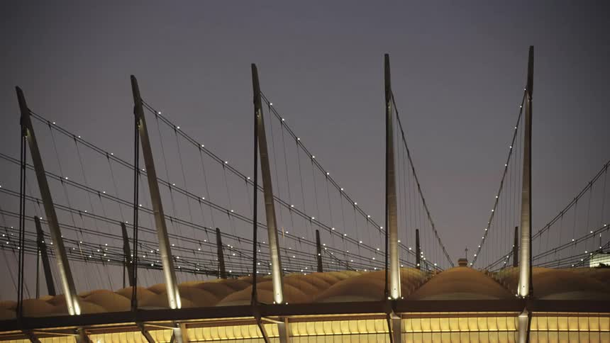A modern stadiums illuminated roof and striking cable stayed structure stand out against the dark evening sky