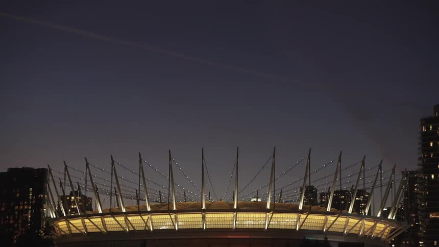 A wide lowangle shot shows a large stadium with bright yellow lights illuminating its cablesupported roof structure surrounded by a city skyline at dusk under a dark blue sky