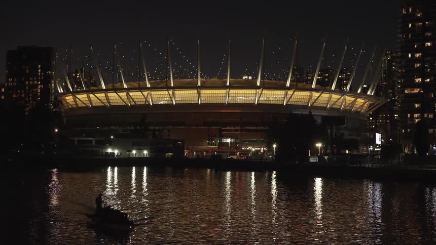 A large brightly lit stadium with a cablesupported roof stands at night along a waterfront with a small boat and the light reflections of surrounding city buildings visible on the dark rippling water