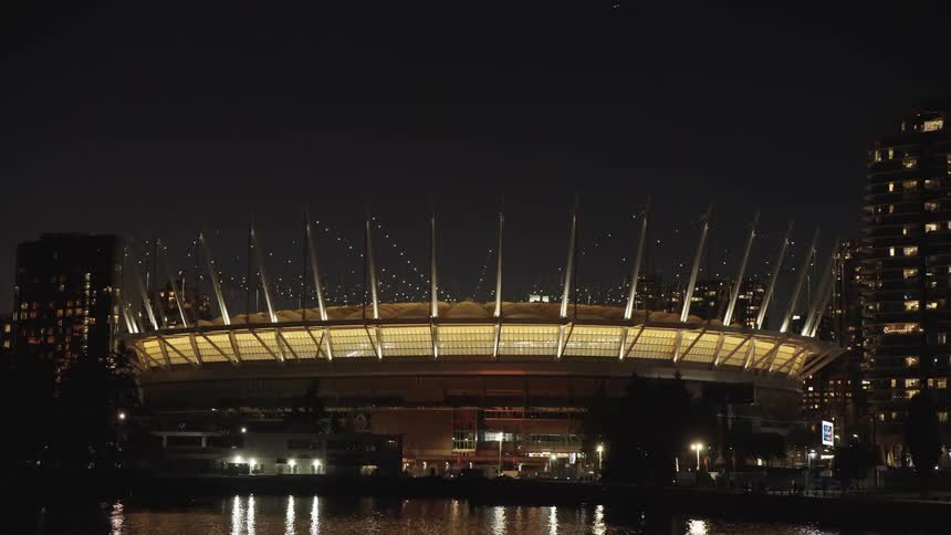 Illuminated stadium stands out at night surrounded by buildings and reflections on the water