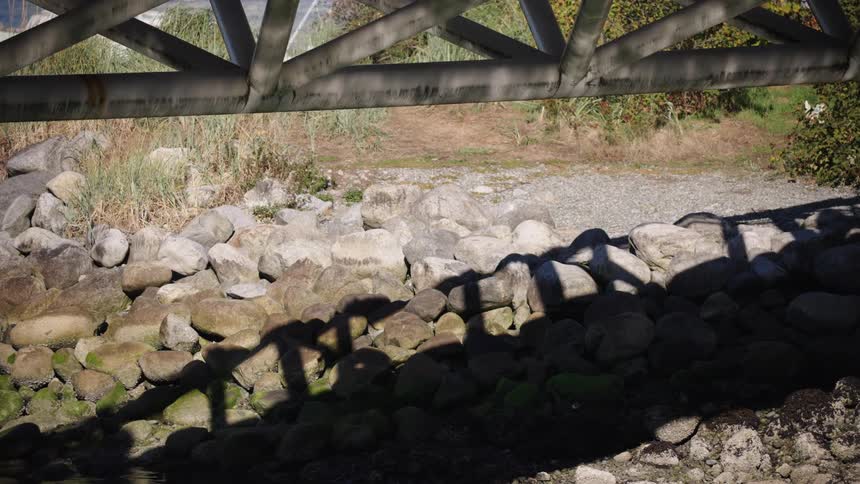 Medium view looking underneath a metal truss structure with sunlight casting the distinct long shadows of multiple unseen figures onto a pile of large foreground rocks