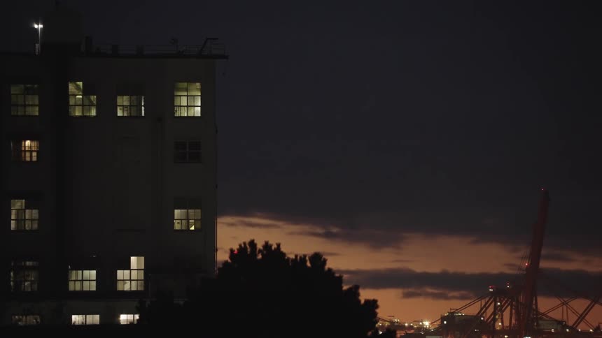 A mediumsized building with lit windows stands in the dark against a cloudy evening sky with orange light near the horizon and a large industrial crane visible in the bottom right corner