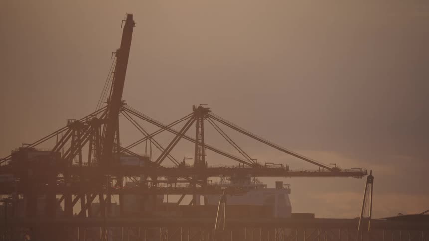 Large intricate industrial machinery and a massive vessel silhouetted against a pale orange hued sky with a bird flying across the upper frame