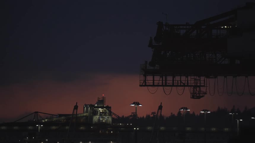 This is a lowlight distant shot of an industrial dock or facility with large machinery silhouetted against a dark dramatic twilight skythe scene is lit by scattered bright white lamps illuminating the foreground and creating a stark contras