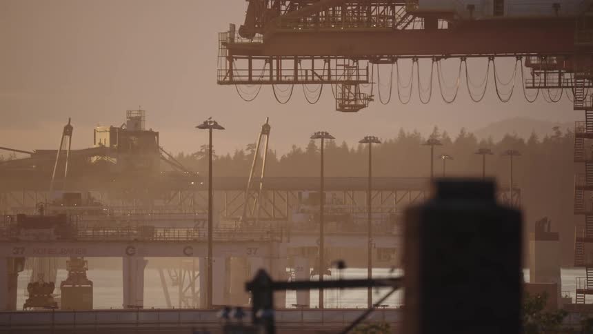 A hazy telephoto view shows the silhouette of massive shipping cranes and industrial buildings at a busy port with the sun low on the horizon