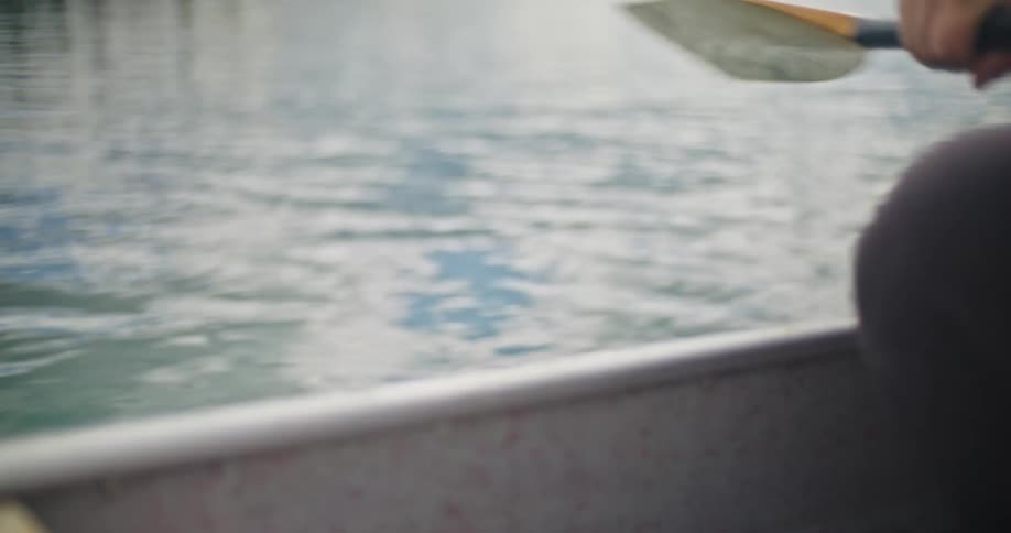 Inside a boats rim repeatedly showing a persons hand lowering a paddle with a colored blade into the water and lifting it out