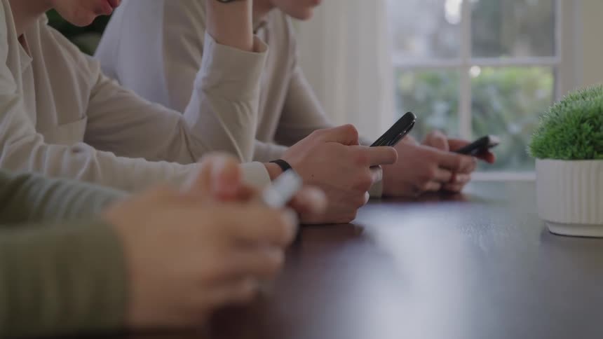 Three people seated at a table each holding and looking at a cellphone with a potted plant and a sunlit window in the background