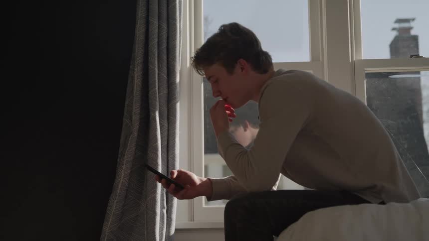 A man seated on a bed looking at a cellphone held in his hand positioned in front of a bright window with curtains hanging on the side