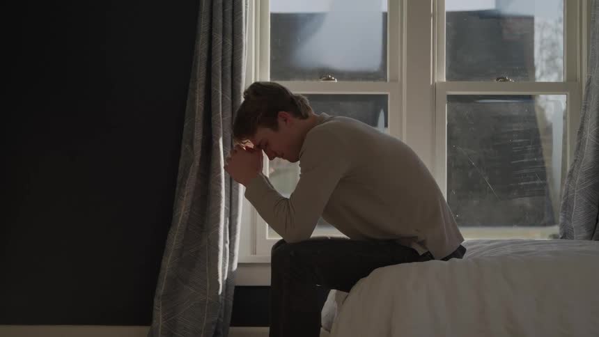 A man sitting on the edge of a bed with his head resting in his hands positioned against a large bright window in a room with a darkpainted wall