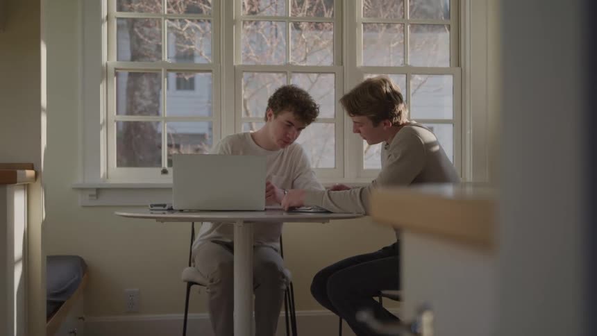 Two men work together on a laptop and papers at a table indoors