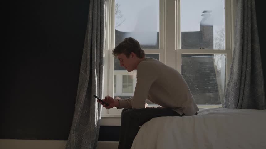 A young man sits on the edge of a bed by a sunlit window checking his phone and looking thoughtful
