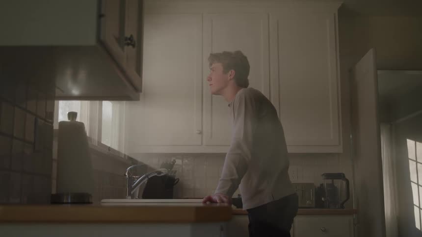 A young man leans on a kitchen counter gazing out the window with a thoughtful slightly concerned expression
