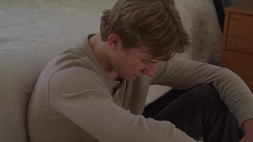A young man with blonde hair wearing a light gray longsleeve shirt sits slumped against a wall on the floor of a bedroom looking down with an expression of distress or sadness