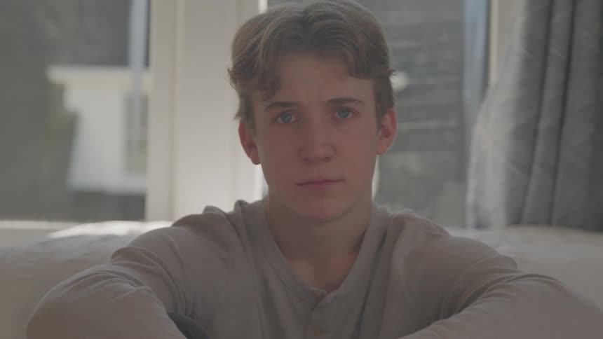 This is a medium closeup shot of a young man with light brown hair sitting indoors against a brightly lit windowhe is initially looking down and then looks directly at the camera with a calm neutral expression