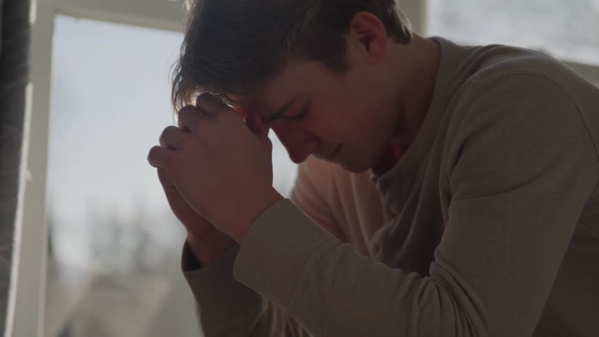Close up of a young man sitting contemplatively with his head resting on his hands in a room during daytime
