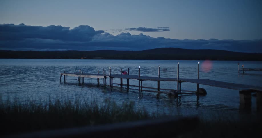 A long wooden dock with small lights extends into a dark lake with rolling hills visible on the opposite shore under a dramatic cloudy blue sky at dusk