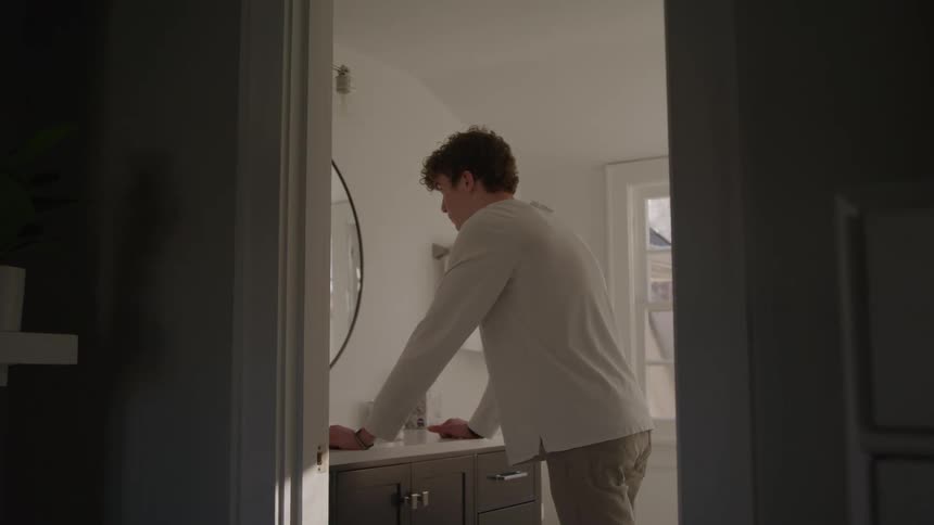 A young man with curly hair wearing a light gray longsleeve shirt and khaki pants leans over a bathroom vanity with a round mirror seen through a dark door frame
