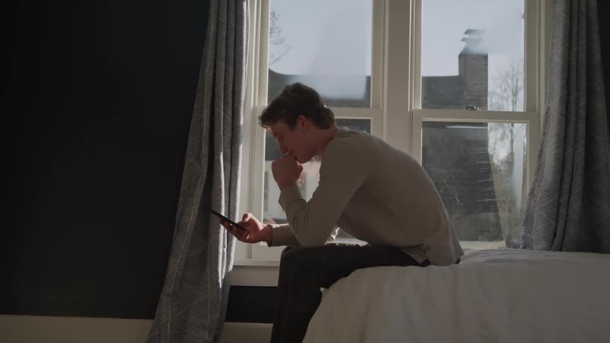 A young man sits contemplatively on the edge of a bed looking down at his smartphone near a bright window