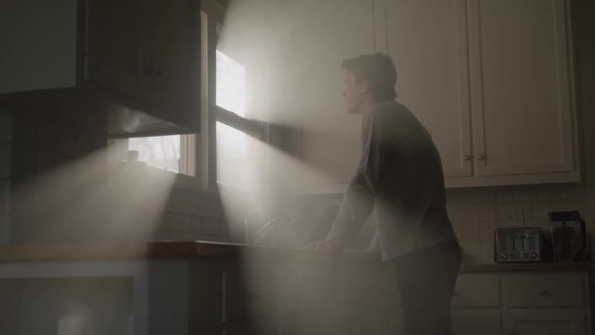 Sunbeams slice through a kitchen window as a young man leans over the counter looking thoughtful and tense
