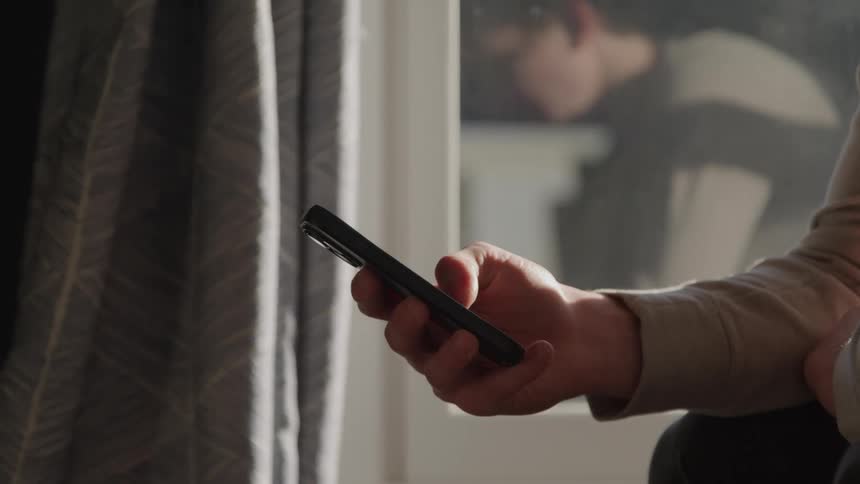 A human hand holds a mobile phone next to a window with closed curtains inside a structure