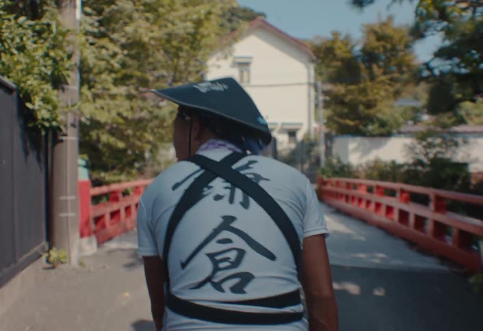 View from behind of a man wearing a harness and traditional hat walking away from the camera moving across a small redrailed bridge toward a white house surrounded by green trees