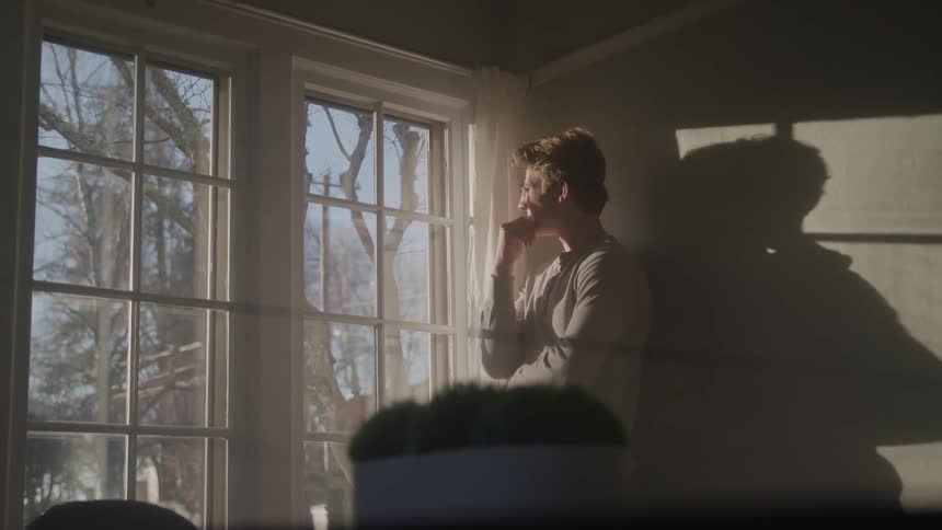 A young man gazes out the window on a sunny day his face reflecting sadness
