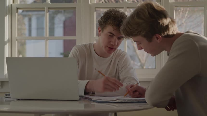 Two young men study together at a table with notebooks and a laptop