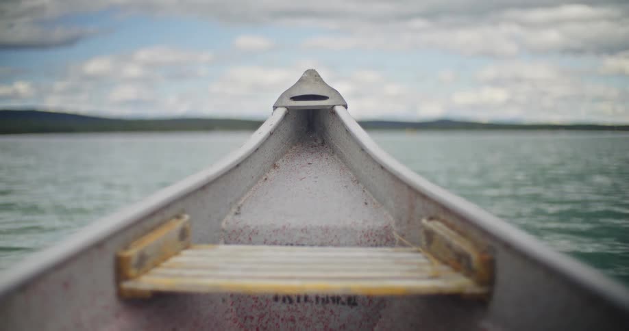 The front of a vessel floating on water moving toward a distant shoreline lined with trees and residential structures