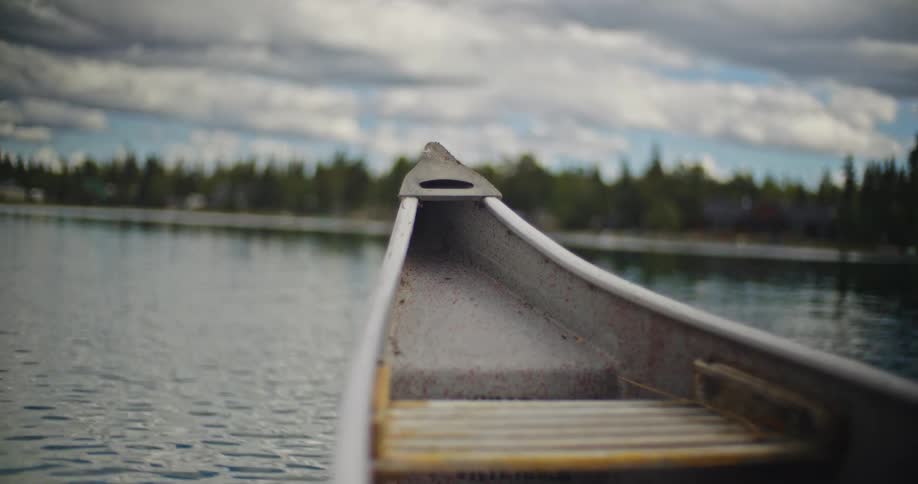 The bow of a vessel floating on a body of water facing a distant shoreline of dense trees under a sky filled with white and gray clouds