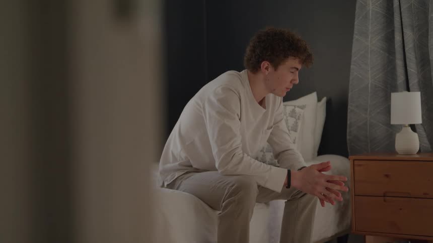 A young man sits on his bed in a bedroom at midday his expression marked by sadness