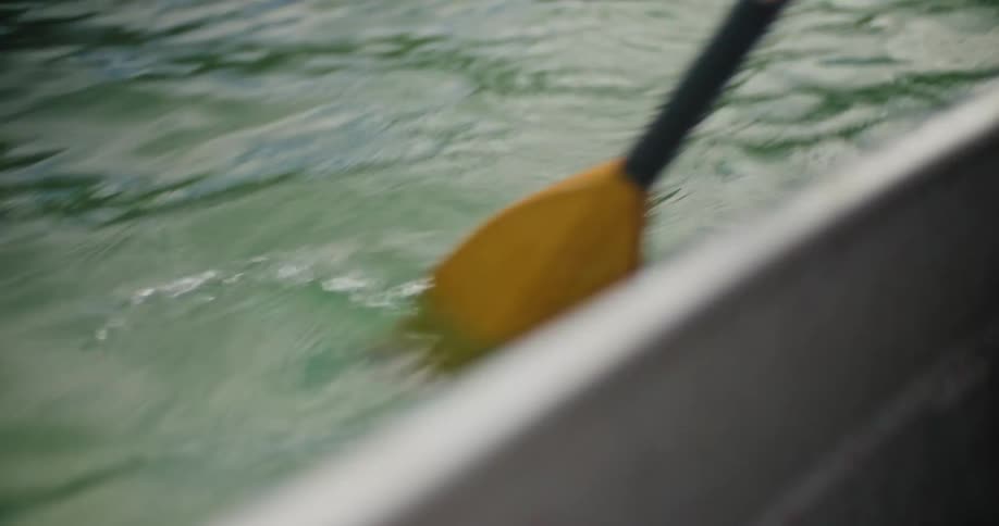 A yellow and black paddle repeatedly dips into and pulls through the clear green water beside the gray edge of a floating vessel