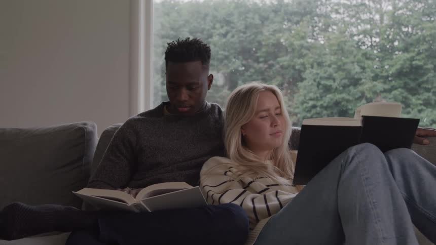 A couple sits on a couch reading by a window in a living room