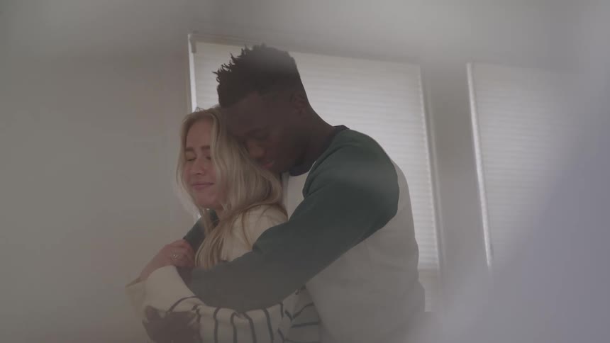 A couple standing and holding each other close in front of a window with blinds illuminated by bright light from behind