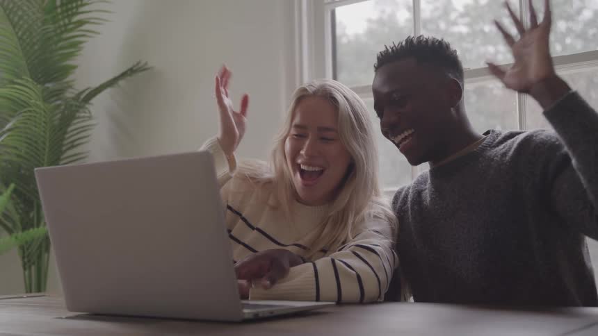 A black man and a blonde woman in sweaters look at a laptop screen and raise their hands in excitement celebrating good news in front of a bright window