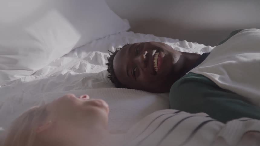 A happy couple lies in bed laughing and enjoying a sunlit morning together
