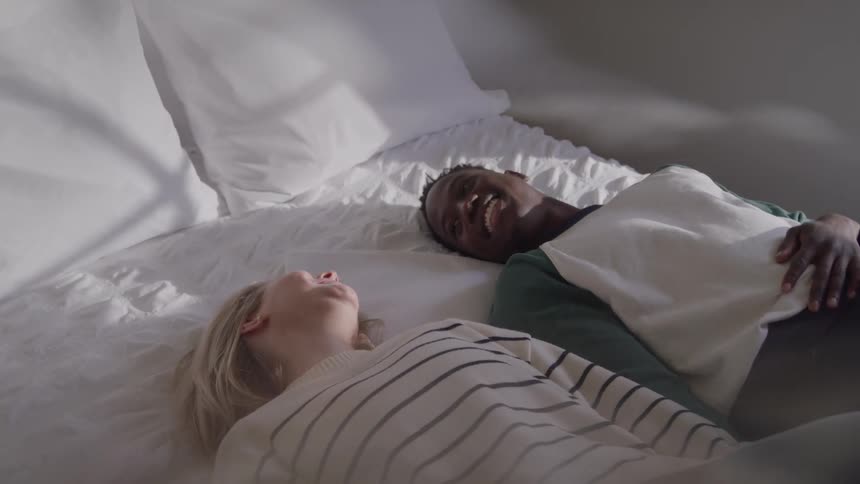 A blond woman in a striped shirt and a man in a green shirt and white sweater are lying on a bed with white bedding laughing and reaching their arms up in a bright bedroom