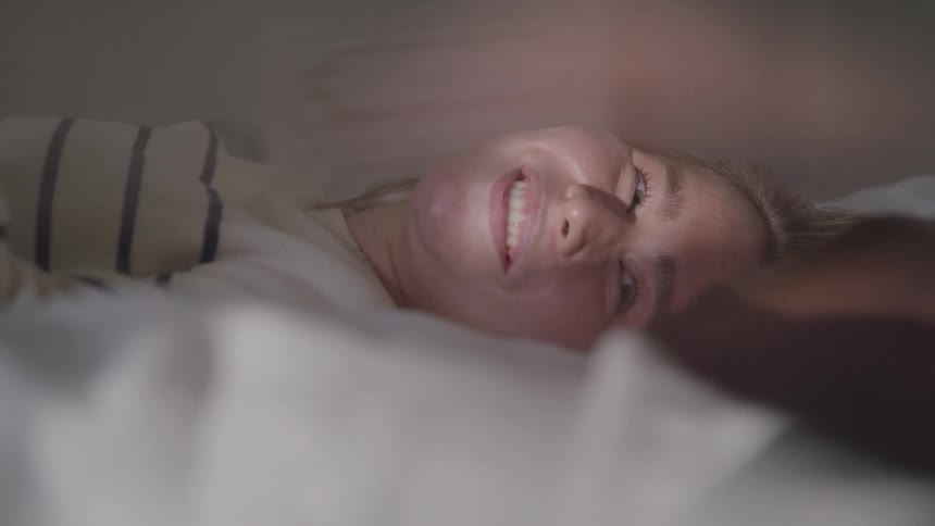 A young woman with blonde hair lies on her side in a bed with white pillows and blankets smiling happily as light illuminates her face from the side